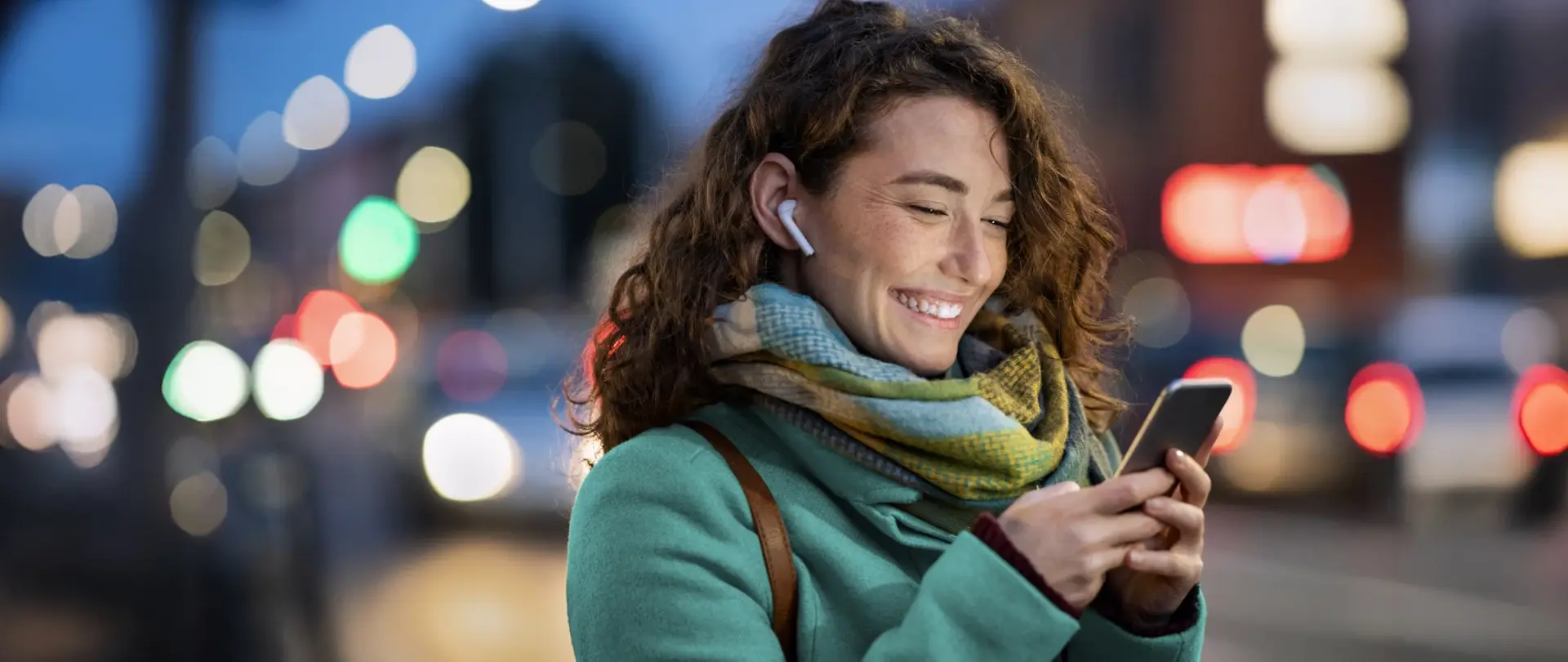 Eine Person mit Smartphone und Kopfhörern steht abends in einer beleuchteten Stadtumgebung mit verschwommenem Straßenhintergrund.