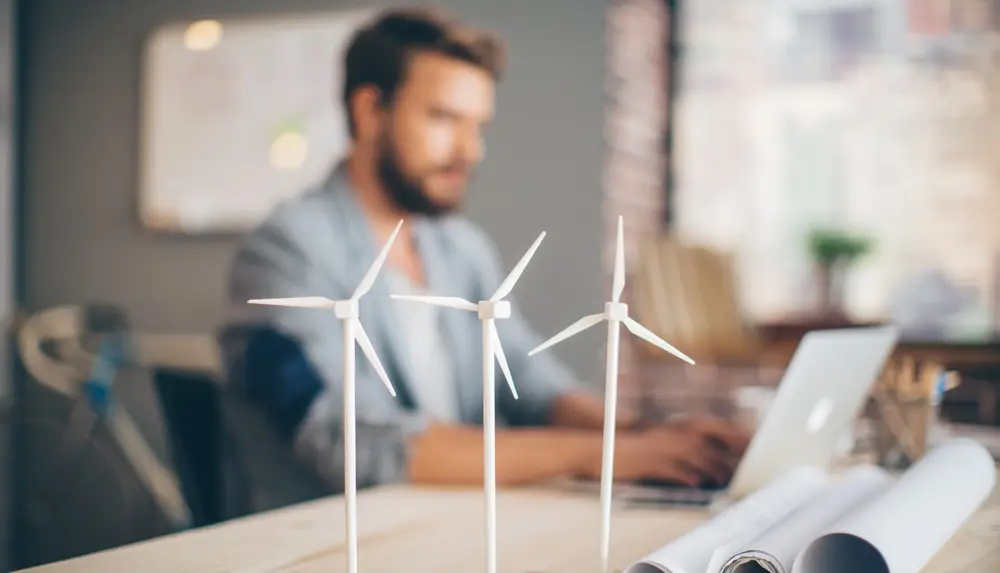 Ein Mann sitzt an seinem Schreibtisch und plant ein Windkraftprojekt, während im Vordergrund auf dem Tisch drei Windkrafträdermodelle stehen.