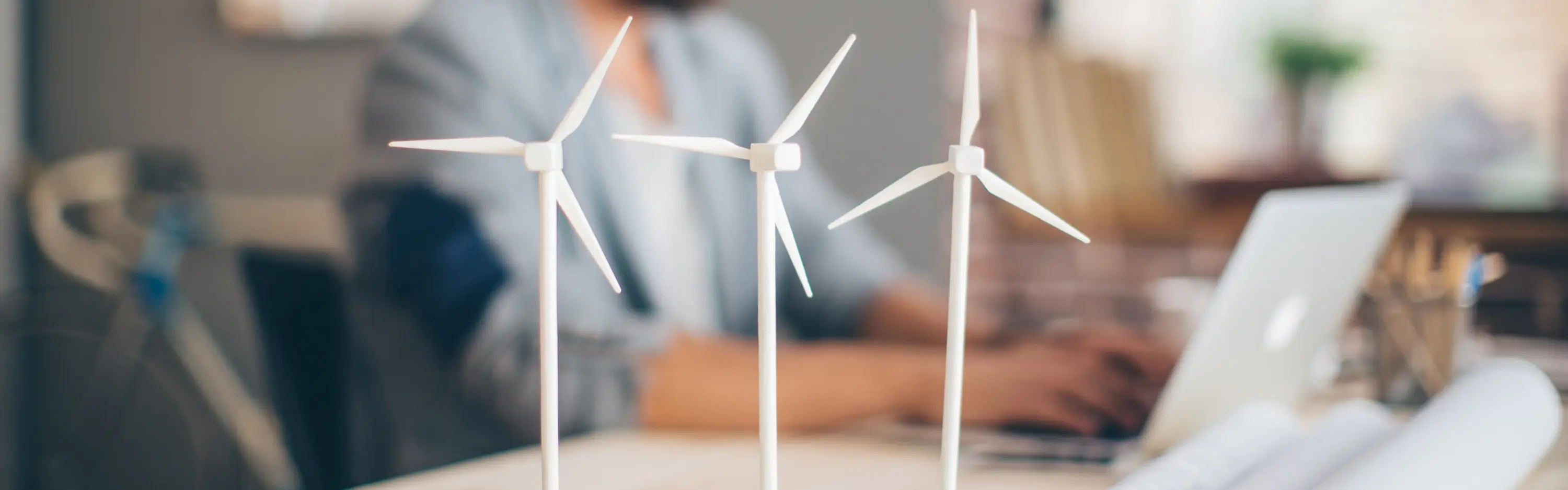 Ein Mann sitzt an seinem Schreibtisch und plant ein Windkraftprojekt, während im Vordergrund auf dem Tisch drei Windkrafträdermodelle stehen.