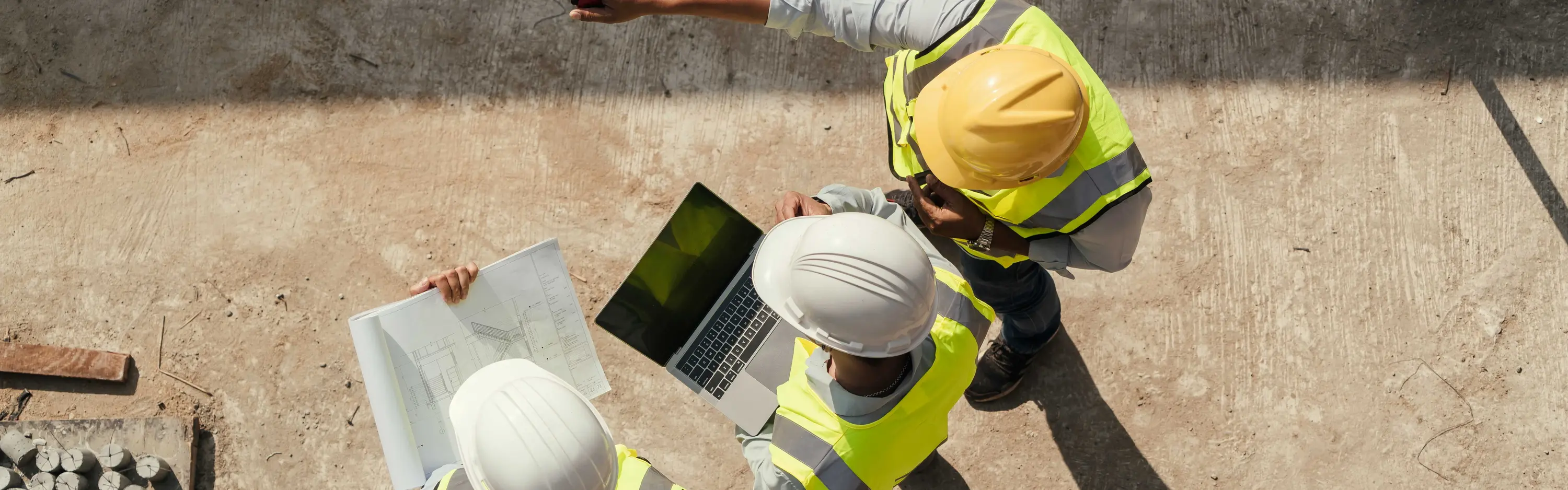 Drei Personen mit Schutzkleidung und Plänen sowie Laptop befinden sich auf einer Baustelle und wurden aus der Vogelperspektive fotografiert.