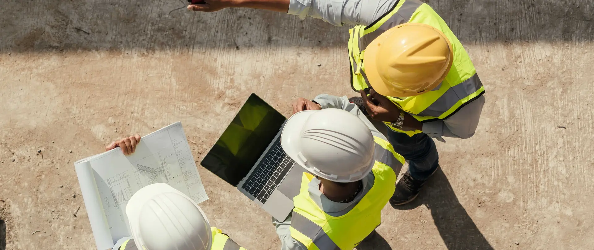 Drei Personen mit Schutzkleidung und Plänen sowie Laptop befinden sich auf einer Baustelle und wurden aus der Vogelperspektive fotografiert.
