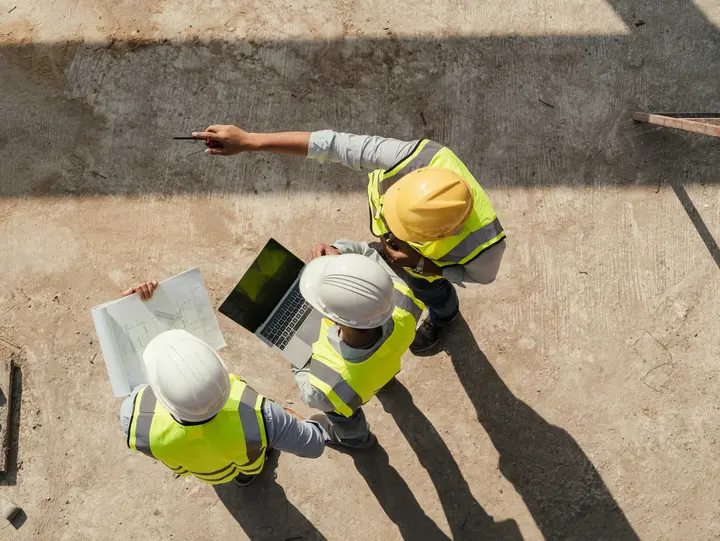Drei Personen mit Schutzkleidung und Plänen sowie Laptop befinden sich auf einer Baustelle und wurden aus der Vogelperspektive fotografiert.