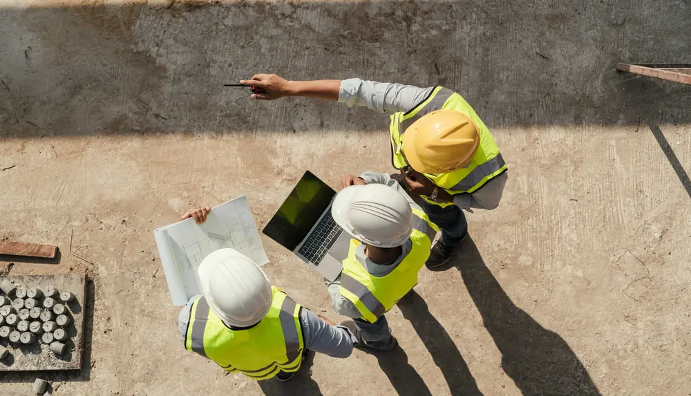 Drei Personen mit Schutzkleidung und Plänen sowie Laptop befinden sich auf einer Baustelle und wurden aus der Vogelperspektive fotografiert.