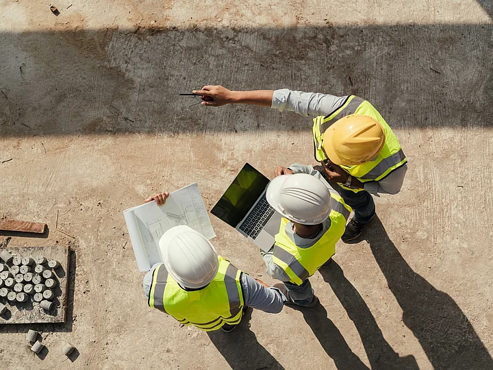 Professioneller Kabel- und Rohrleitungsbau Drei Personen mit Schutzkleidung und Plänen sowie Laptop befinden sich auf einer Baustelle und wurden aus der Vogelperspektive fotografiert.
