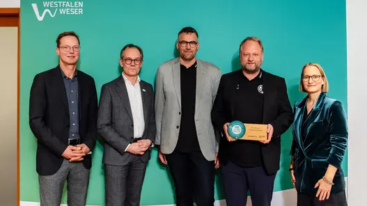 Eine Gruppe aus vier Männern und einer Frau stehen vor einer petrolfarbenen Werbewand von Westfalen Weser. Ein Mann im Jackett hält den Preis in den Händen.