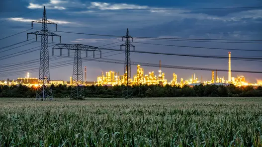 Strommasten und Hochspannungsleitungen führen zu einer beleuchteten Industrieanlage bei Dämmerung.