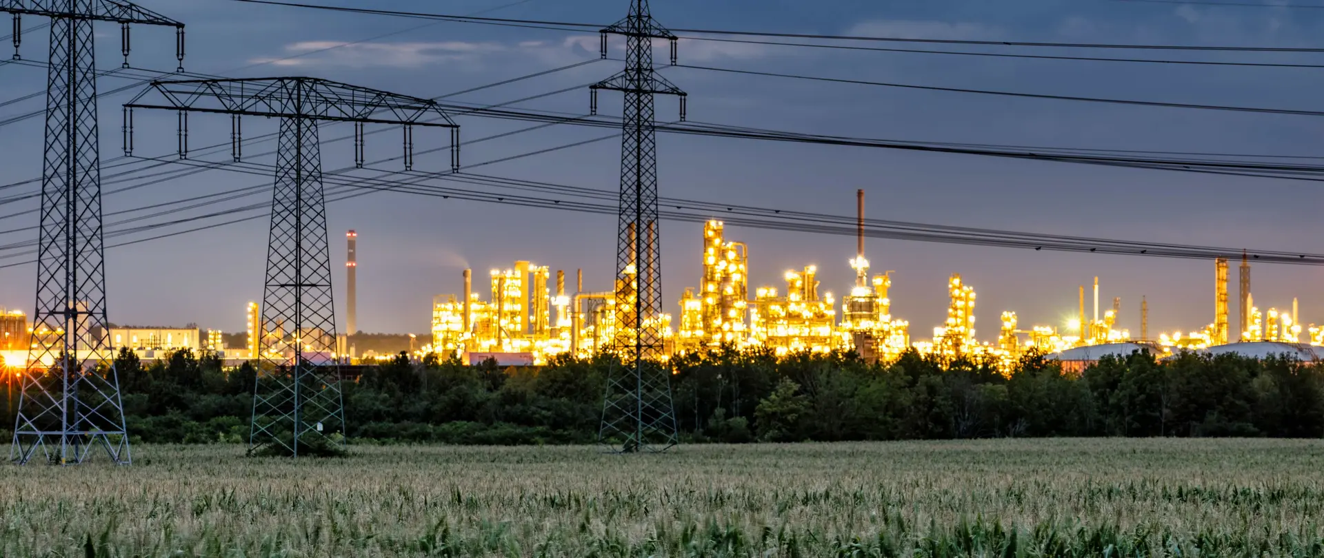 Strommasten und Hochspannungsleitungen führen zu einer beleuchteten Industrieanlage bei Dämmerung.