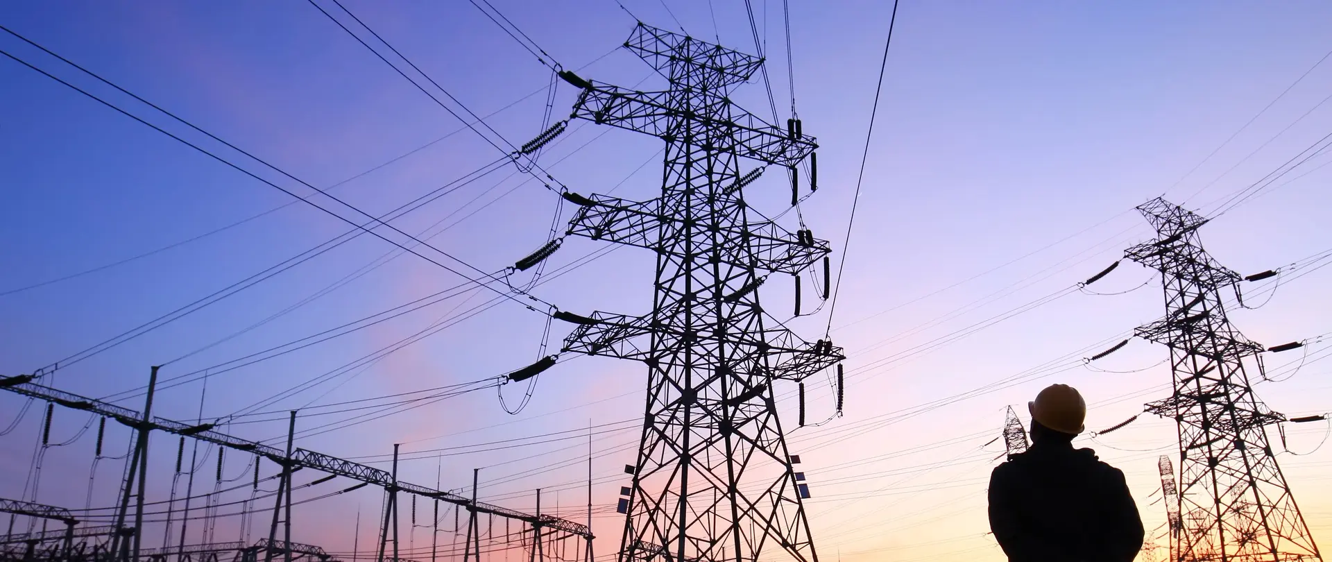 Ein Mann steht bei Sonnenuntergang vor einem großen Umspannwerk, während der Himmel lila und pink leuchtet.