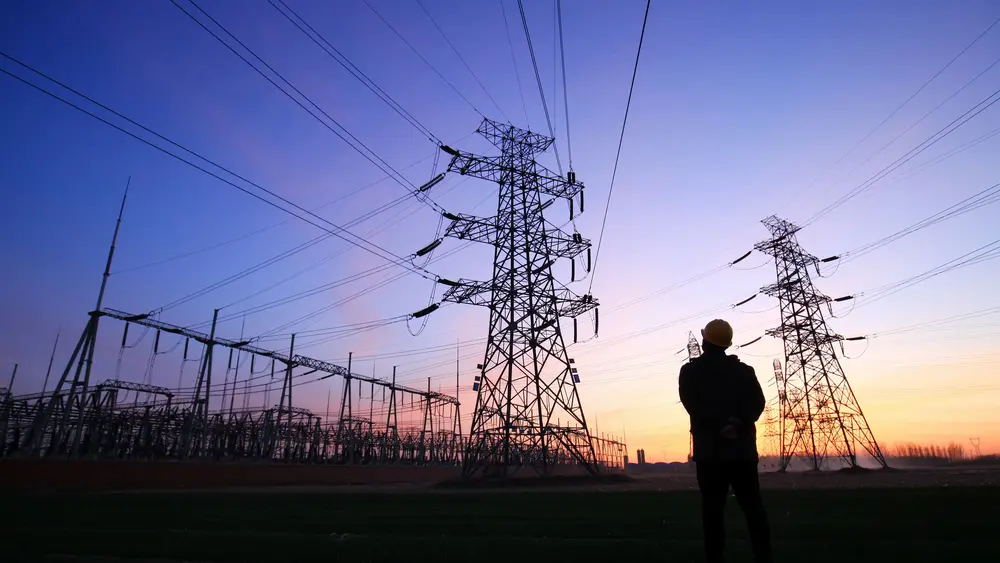Ein Mann steht bei Sonnenuntergang vor einem großen Umspannwerk, während der Himmel lila und pink leuchtet.