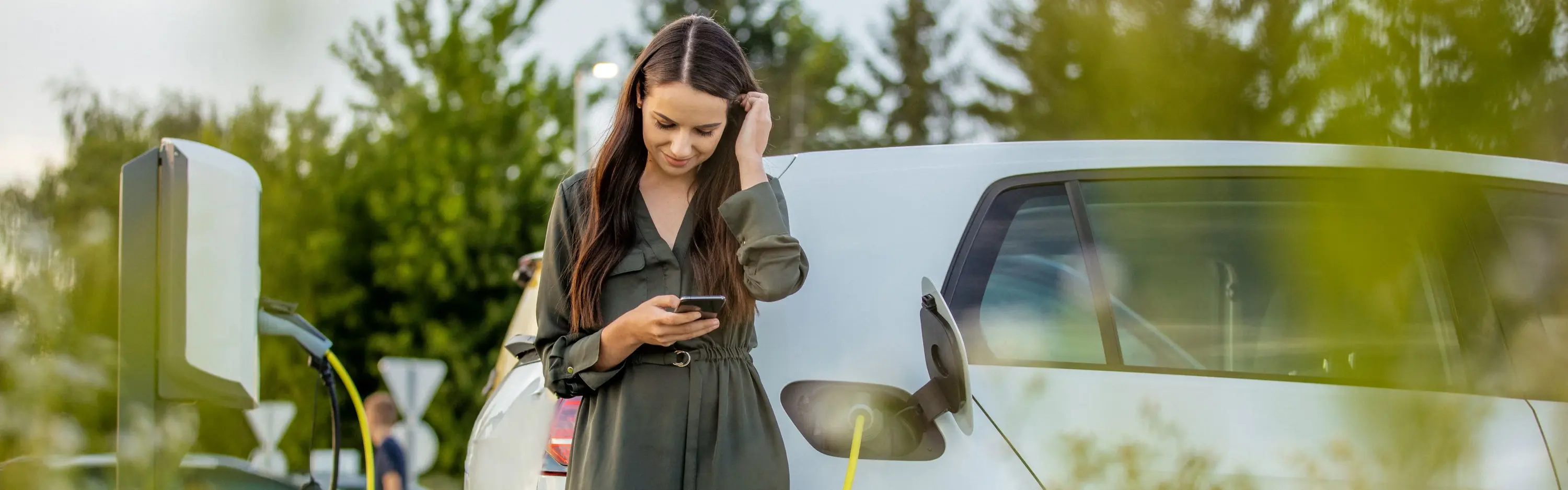 Eine Person lädt ein weißes Elektroauto an einer Wallbox und schaut dabei auf ihr Smartphone.