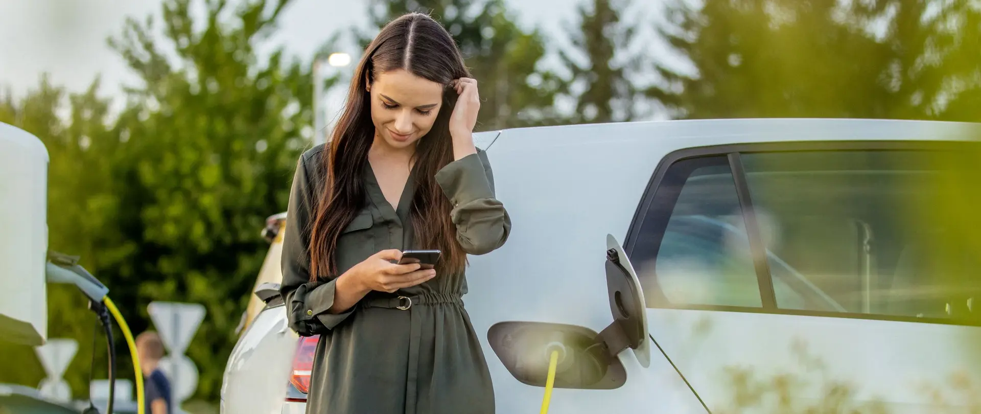 Eine Person lädt ein weißes Elektroauto an einer Wallbox und schaut dabei auf ihr Smartphone.