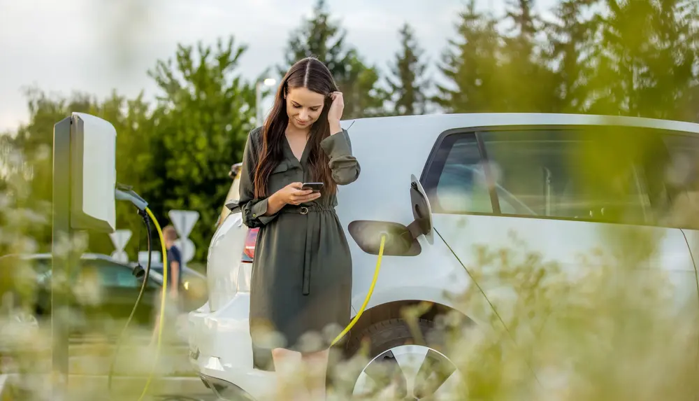 Eine Person lädt ein weißes Elektroauto an einer Wallbox und schaut dabei auf ihr Smartphone.