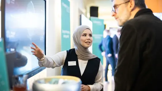 Westfalen Weser Forum Projektvorstellung Eine junge Frau erklärt an einem großen Bildschirm das Projekt Energiekompass Im Vordergrund sieht man einen Mann, der interessiert zuhört.