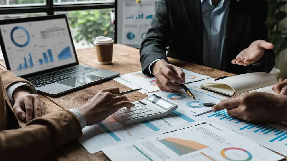 Drei Personen sitzen an einem Tisch, auf dem Blätter mit verschiedenen Statistiken liegen ebenso wie ein Laptop.