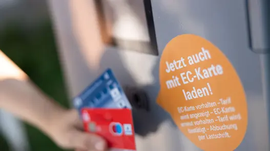 Eine Person hält zwei Karten vor eine Ladesäule mit Hinweis zur EC-Karten-Zahlung per Lastschrift.