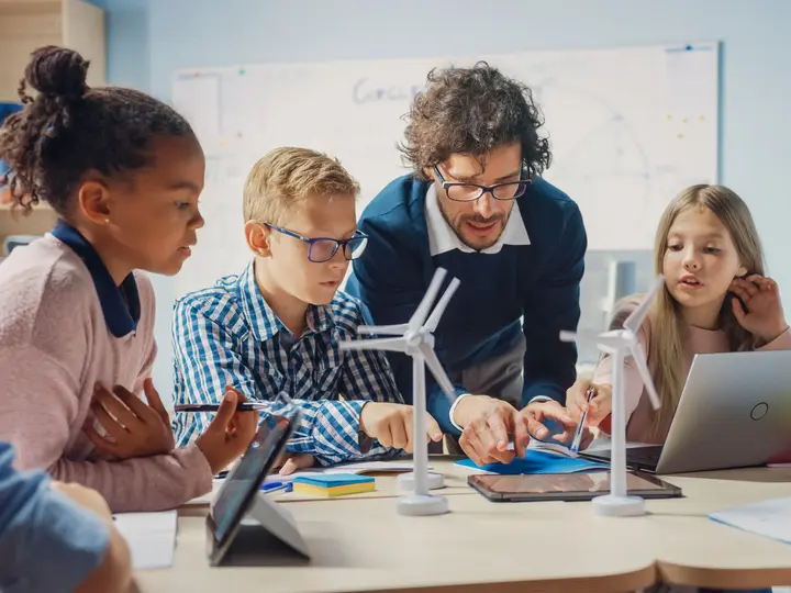 Kinder und eine erwachsene Person experimentieren gemeinsam mit Windrad-Modellen und digitalen Geräten in einem modernen Klassenraum.
