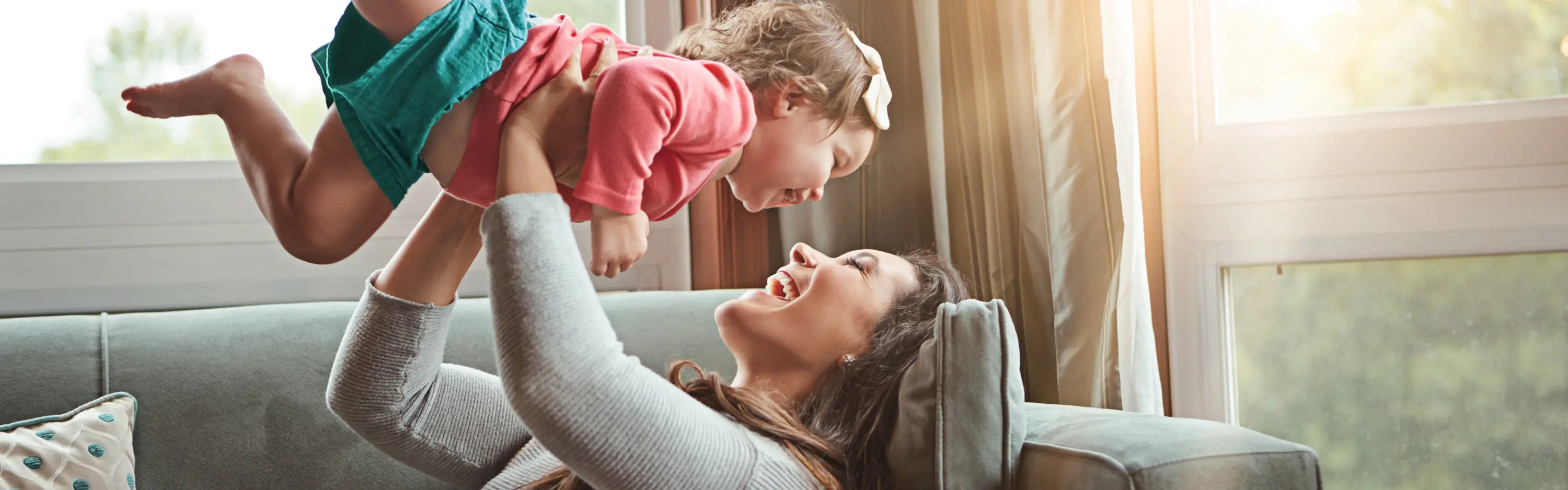 Eine Mutter liegt auf dem Sofa in einem gemütlichem Wohnzimmer. Die Mutter spielt mit der Tochter und beide sind sehr glücklich, Die Sonne scheint durch die großen Fenster rein,