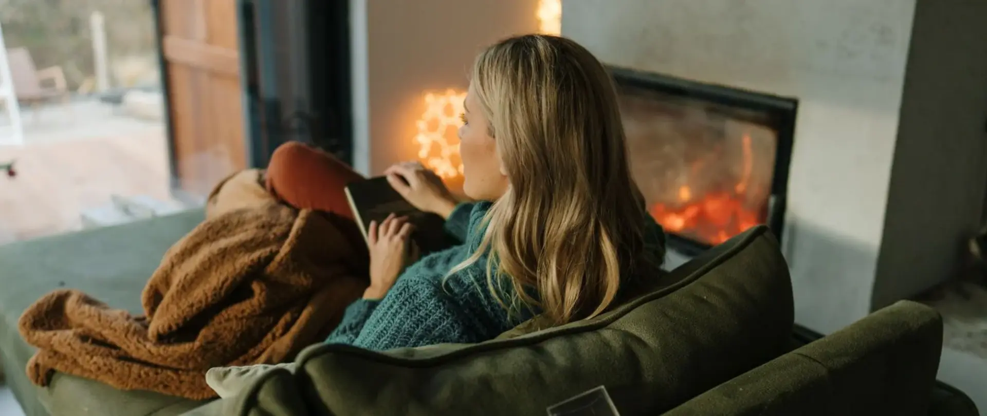 Eine Frau sitzt auf der Couch unter einer Decke. Sie hat ein Buch auf ihrem Schoß und schaut aus dem Fenster. Der Kamin ist an