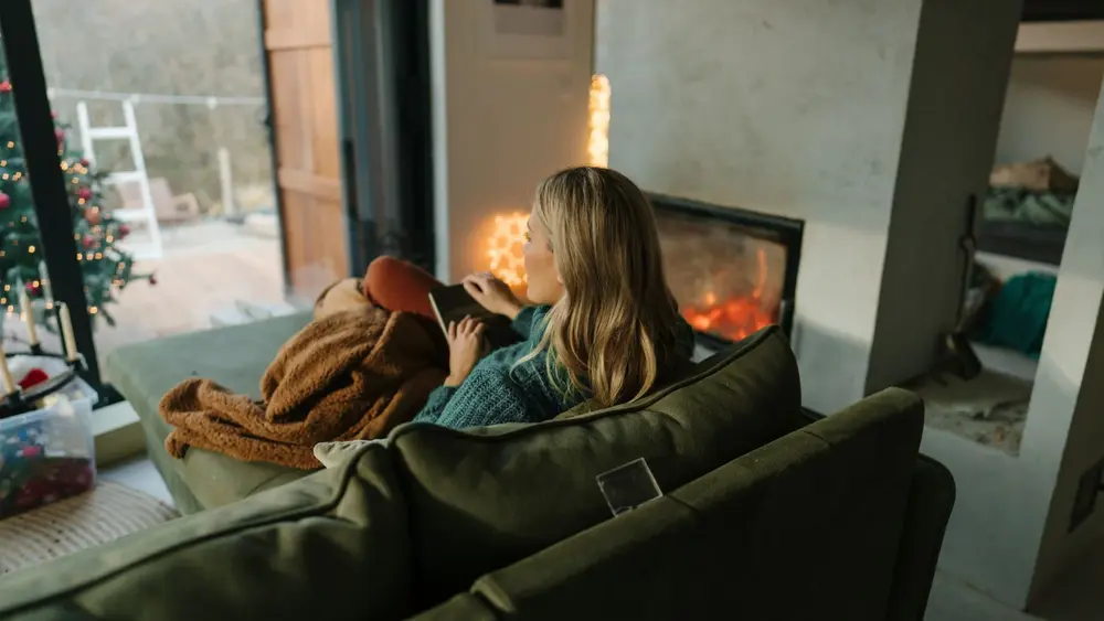 Eine Frau sitzt auf der Couch unter einer Decke. Sie hat ein Buch auf ihrem Schoß und schaut aus dem Fenster. Der Kamin ist an