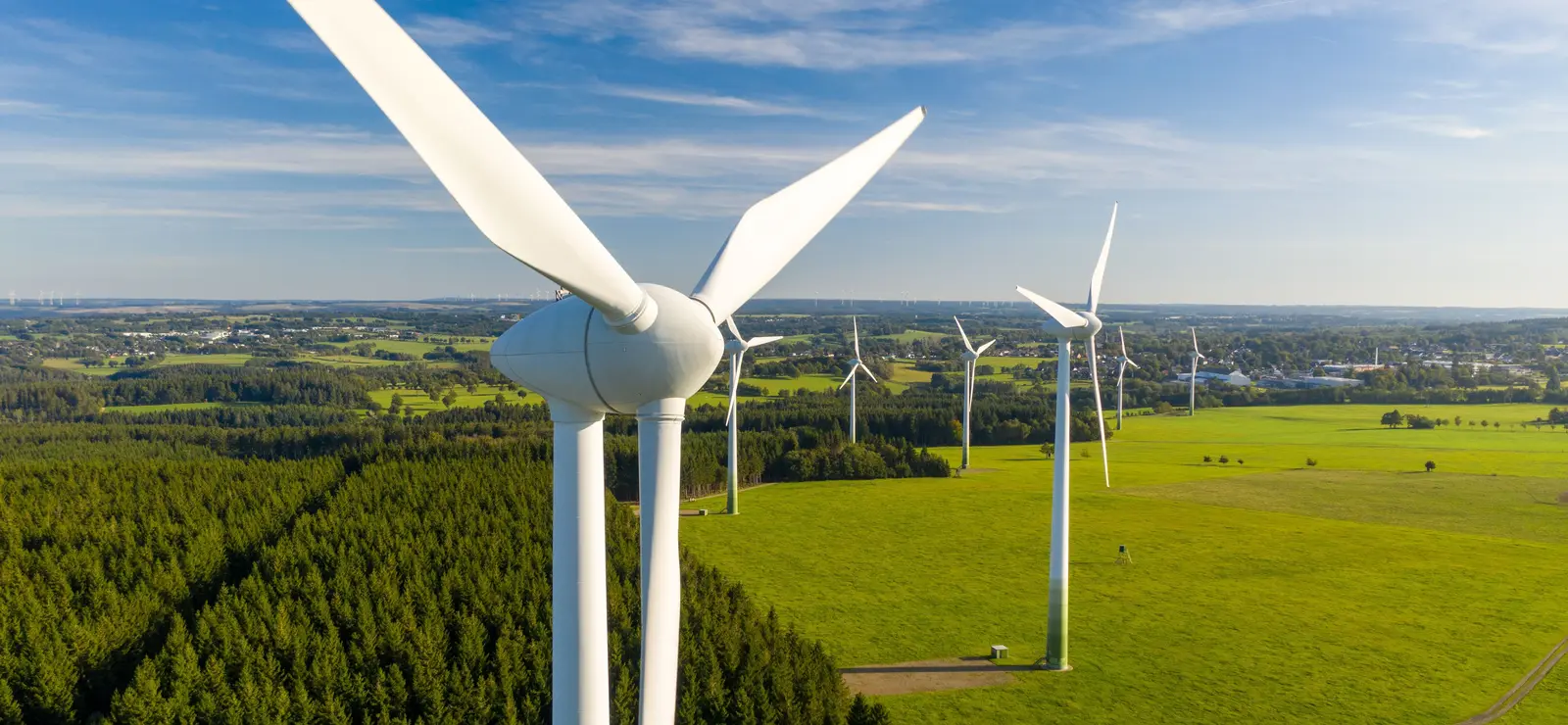 Mehrere Windkraftanlagen stehen auf grünen Wiesen unter blauem Himmel mit vereinzelten Wolken.