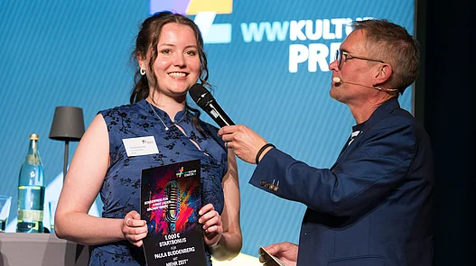 Paula Buddenberg hält eine Urkunde für den Start-Bonus des WWKulturSTARTER26 in der Hand, während der Moderator Ingo Börchers ein Interview auf der Bühne führt.