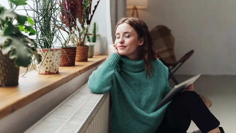 Frau sitzt im beheizten Wohnzimmer und lehnt auf der Fensterbank