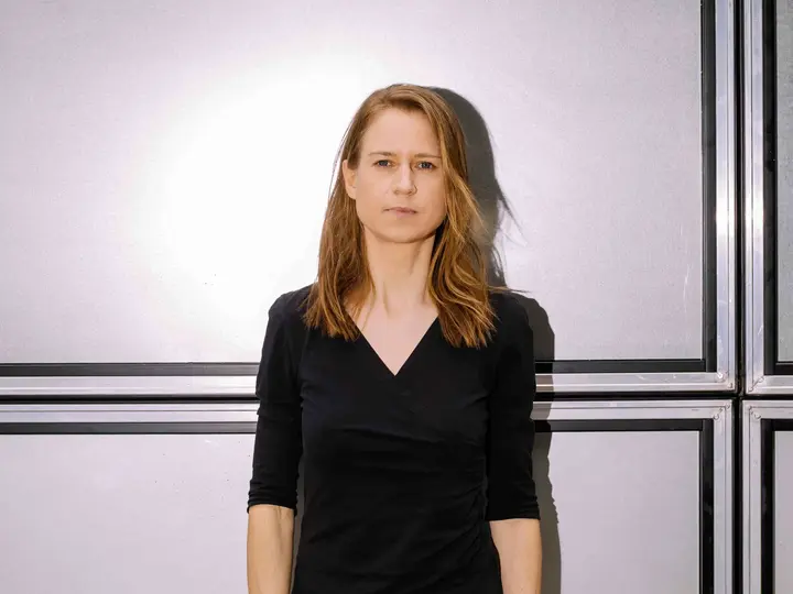 Eine Frau in schwarzer Kleidung und mittellangen Haaren steht vor einer hellen Wand.
