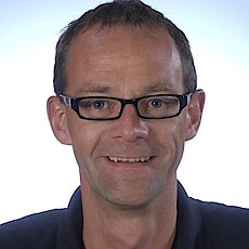 Portraitfoto einer männliche Person mit Brille und dunkelblauen Oberteil auf weißem Hintergrund 