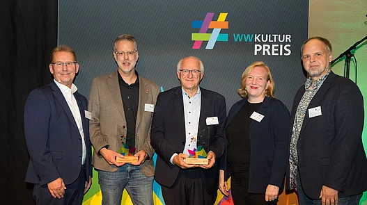 Platz 1: KZ-Gedenk-und Dokumentationsstätte Porta Westfalica Gruppenfoto vor einer Fotowand von Vereinsmitgliedern der Gedenk- und Dokumentationsstätte Porta Westfalica mit dem 1. Hauptpreis des WWKulturpreis26..
