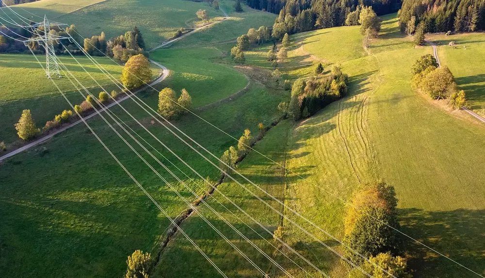 Das Bild zeigt Stromleitungen, die sich durch eine grüne, ländliche Landschaft mit Wiesen und Hügeln ziehen.