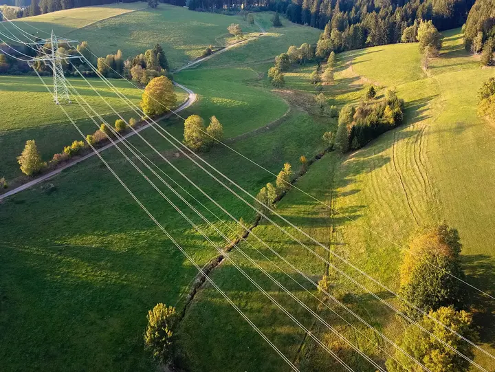 Das Bild zeigt Stromleitungen, die sich durch eine grüne, ländliche Landschaft mit Wiesen und Hügeln ziehen.