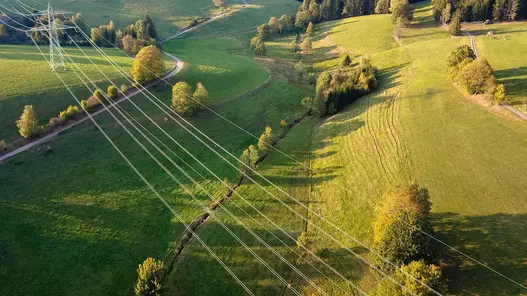 Das Bild zeigt Stromleitungen, die sich durch eine grüne, ländliche Landschaft mit Wiesen und Hügeln ziehen.