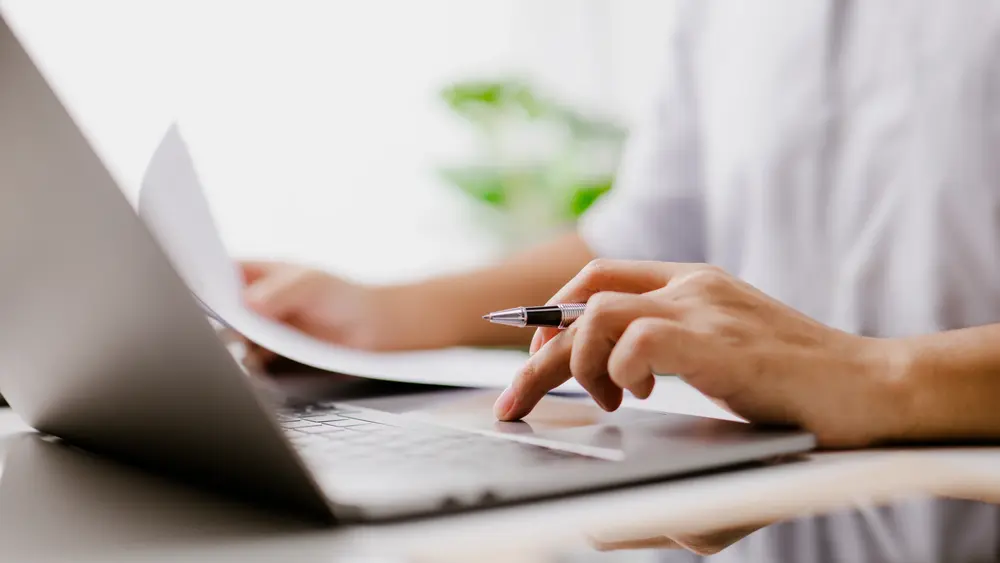 Eine Person tippt auf ihrem Laptop Daten ein und hat weitere Unterlagen in Papierform auf dem Tisch vor sich liegen.