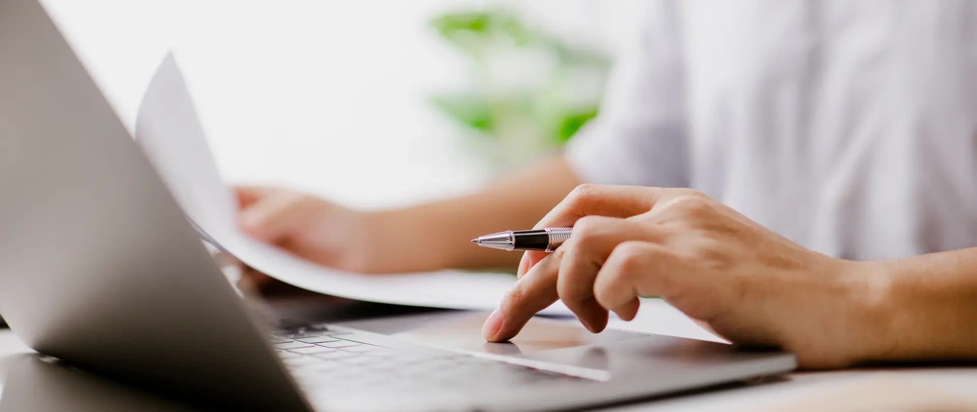 Eine Person tippt auf ihrem Laptop Daten ein und hat weitere Unterlagen in Papierform auf dem Tisch vor sich liegen.