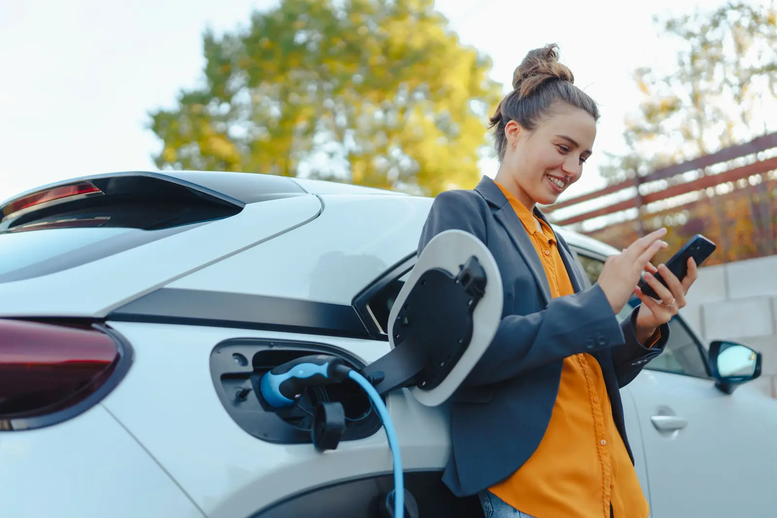 Eine Person steht neben einem weißen Elektroauto, das geladen wird, und nutzt dabei ein Smartphone.