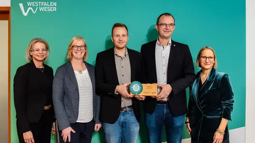 Fünf Personen stehen in einer Reihe vor einer großen Westfalen Weser Wand. Zwei davon halten die Auszeichnung in den Händen.