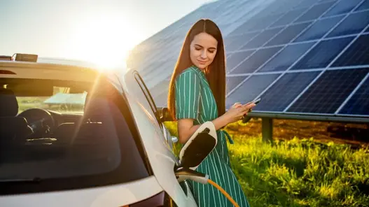 Elektroauto am Ladestecker, Frau wartet glücklich im Sonnenuntergang