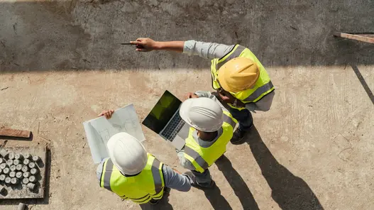 Drei Bauarbeiter mit Helm und Laptop besprechen einen Bauplan auf einer Baustelle.