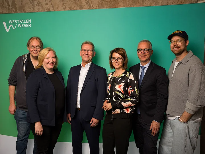 Sechs Mitglieder der Jury vom WWKulturSTARTER26 stehen vor einer grünen Westfalen-Weser-Fotowand.