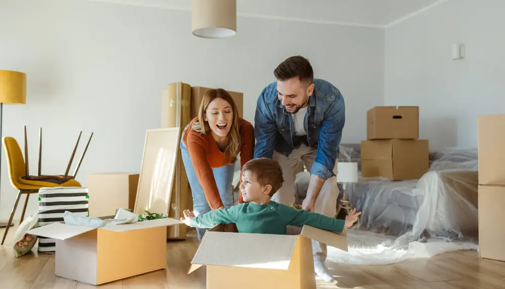 Familie mitten im Umzug Eine Familie mit kleinem Sohn schiebt zwischen Kartons ihren Sohn im Karton durch das neue Wohnzimmer