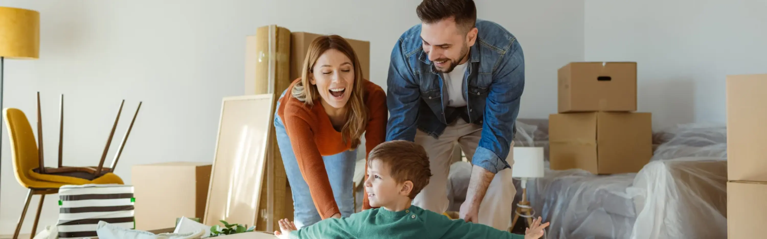 Familie mitten im Umzug Eine Familie mit kleinem Sohn schiebt zwischen Kartons ihren Sohn im Karton durch das neue Wohnzimmer