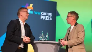 Zwei Männer stehen an einem Stehtisch vor einem großen Banner und unterhalten sich
