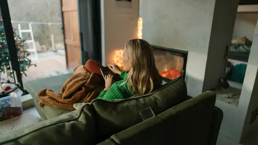Eine Frau sitzt im gemütlichen Wohnzimmer auf ihrem Sofa und schaut nach draußen. Der Kamin ist an und es ist ein kleiner Teil eines Weihnachtsbaums zu sehen