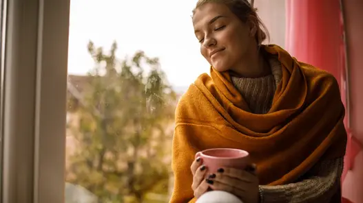 Eine in der Decke eingewickelte Frau sitzt auf einer Fensterbank, hält eine Tasse Kaffee in den Händen und genießt den Moment. Ihre Augen sind geschlossen.