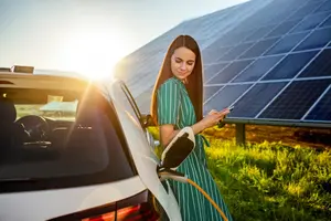 Eine Frau steht angelehnt an einem Elektro-Auto und hat ein Handy in der Hand. Das Auto steht neben einer großen Photovoltaik-Anlage. Sie Sonne geht gerade unter