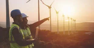 Zwei Personen mit Schutzhelmen zeigen auf Windkraftanlagen in hügeliger Landschaft bei Sonnenuntergang.