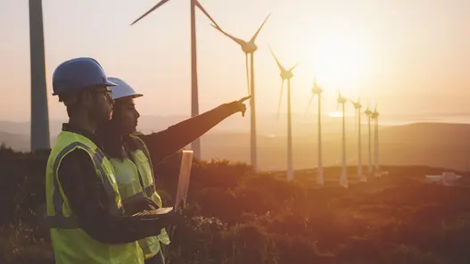 Zwei Personen mit Schutzhelmen zeigen auf Windkraftanlagen in hügeliger Landschaft bei Sonnenuntergang.