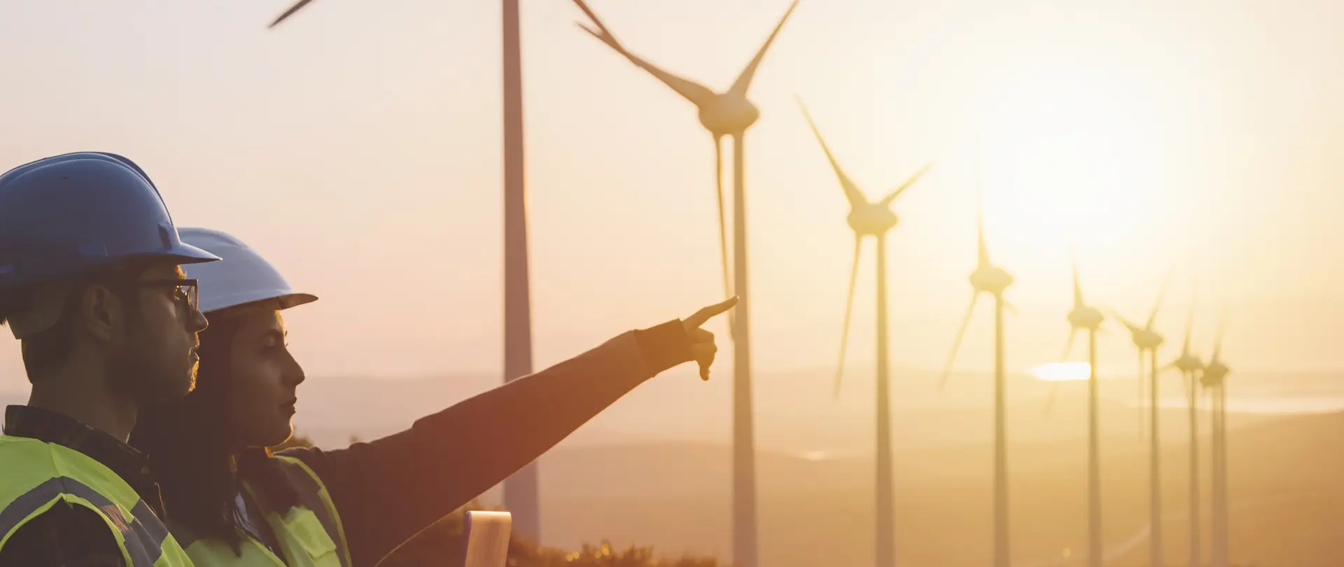 Zwei Personen mit Schutzhelmen zeigen auf Windkraftanlagen in hügeliger Landschaft bei Sonnenuntergang.