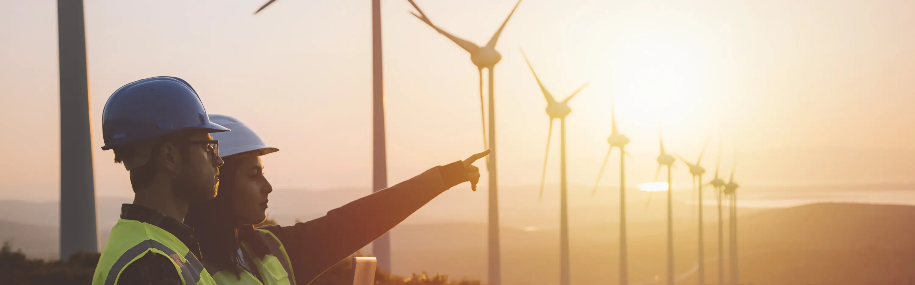 Zwei Personen mit Schutzhelmen zeigen auf Windkraftanlagen in hügeliger Landschaft bei Sonnenuntergang.