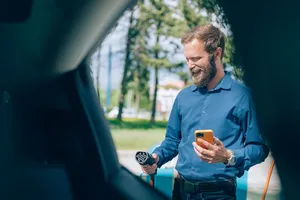 Eine Person steht im Freien und hält ein Ladekabel sowie ein Smartphone neben einer Ladesäule.